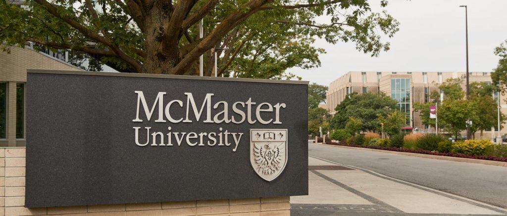 A black stone sign with silver lettering reads “McMaster University”. A silver crest featuring an open book and a winged book also features on the sign. In the background is a street lined with university buildings and foliage.